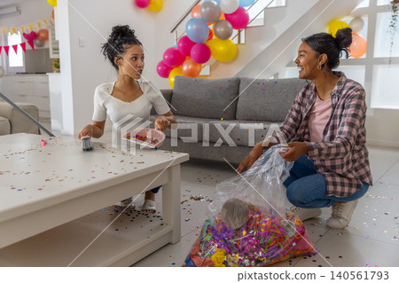 Diverse mom and teenage daughter sweeping confetti into dustpan at home with balloons Diverse mom and teenage daughter sweeping confetti into dustpan at home with balloons 140561793