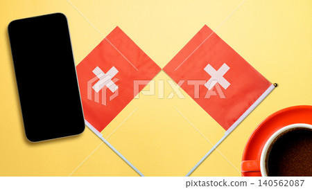 Flat lay composition with two small Swiss flags, a smartphone with a blank screen, and a cup of black coffee on a yellow background. Ideal for Swiss business or travel concepts Flat lay composition with two small Swiss flags, a smartphone with a blank screen, and a cup of black coffee on a yellow background. Ideal for Swiss business or travel concepts 140562087