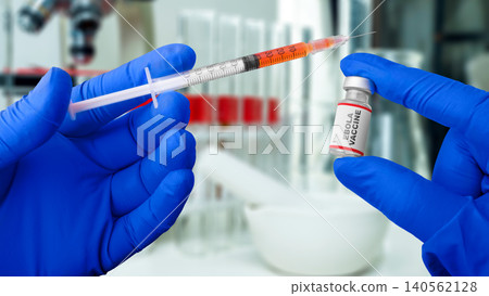 Close-up of a scientist in blue protective gloves holding a syringe with orange fluid and a vial labeled Ebola Vaccine, with blurred laboratory equipment in the background Close-up of a scientist in blue protective gloves holding a syringe with orange fluid and a vial labeled Ebola Vaccine, with blurred laboratory equipment in the background 140562128