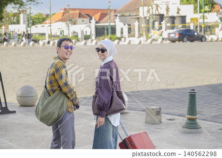 A happy young Indonesian southeast asian muslim couple walks through a sunny city plaza with suitcases, smiling at the camera during their vacation travel. Travel or vacation concept A happy young Indonesian southeast asian muslim couple walks through a sunny city plaza with suitcases, smiling at the camera during their vacation travel. Travel or vacation concept 140562298