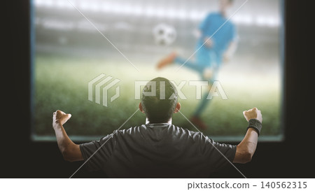 Back view of a passionate sports fan cheering with raised fists while watching a football match on a large television screen at home Back view of a passionate sports fan cheering with raised fists while watching a football match on a large television screen at home 140562315