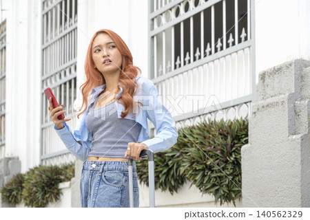 Young Indonesian southeast asian female traveler with red hair looking lost while holding a smartphone and suitcase on a city street. Travel or vacation concept Young Indonesian southeast asian female traveler with red hair looking lost while holding a smartphone and suitcase on a city street. Travel or vacation concept 140562329