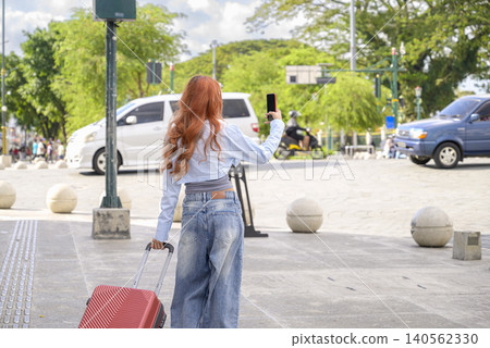Back view of a young Indonesian southeast asian redhead woman holding a red suitcase and taking a selfie with her phone on a sunny city street. Travel or vacation concept Back view of a young Indonesian southeast asian redhead woman holding a red suitcase and taking a selfie with her phone on a sunny city street. Travel or vacation concept 140562330