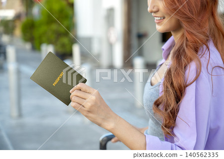 A happy young Indonesian southeast asian female traveler with reddish-brown hair smiles while holding a green passport on a city street, ready for her journey. Travel or vacation concept A happy young Indonesian southeast asian female traveler with reddish-brown hair smiles while holding a green passport on a city street, ready for her journey. Travel or vacation concept 140562335
