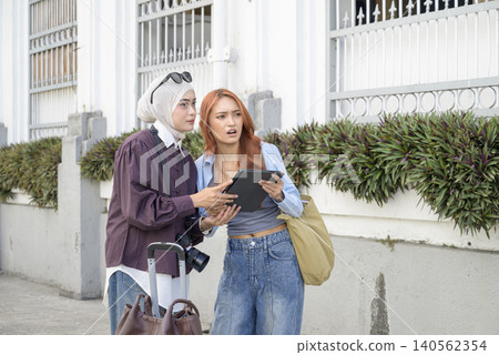Two female Indonesian southeast asian friends, one wearing a hijab, looking confused while navigating city streets using a digital tablet during their travels. Travel or vacation concept Two female Indonesian southeast asian friends, one wearing a hijab, looking confused while navigating city streets using a digital tablet during their travels. Travel or vacation concept 140562354