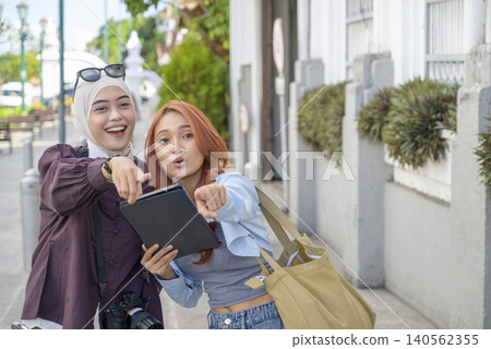 Two happy diverse Indonesian southeast asian women traveling in the city, using a digital tablet and pointing directions while smiling. Travel or vacation concept Two happy diverse Indonesian southeast asian women traveling in the city, using a digital tablet and pointing directions while smiling. Travel or vacation concept 140562355