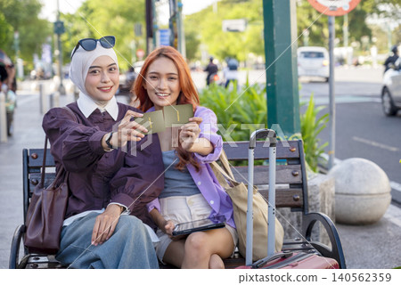 Two young Indonesian southeast asian women, one wearing a hijab, sit on a city bench holding passport and smiling, ready for a vacation trip. Travel or vacation concept Two young Indonesian southeast asian women, one wearing a hijab, sit on a city bench holding passport and smiling, ready for a vacation trip. Travel or vacation concept 140562359
