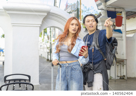 A young Indonesian southeast asian couple traveling together, looking at a smartphone map and pointing directions on an urban street with their luggage. Travel or vacation concept A young Indonesian southeast asian couple traveling together, looking at a smartphone map and pointing directions on an urban street with their luggage. Travel or vacation concept 140562404