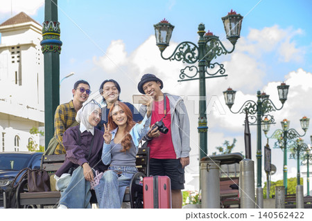 Happy young Indonesian southeast asian tourists take a group selfie with a smartphone while sitting on a street bench during their vacation trip. Travel or vacation concept Happy young Indonesian southeast asian tourists take a group selfie with a smartphone while sitting on a street bench during their vacation trip. Travel or vacation concept 140562422