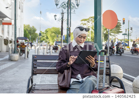 A young Indonesian southeast asian female muslim traveler in a hijab sits on a city bench, using a tablet computer next to her suitcase. Travel or vacation concept A young Indonesian southeast asian female muslim traveler in a hijab sits on a city bench, using a tablet computer next to her suitcase. Travel or vacation concept 140562425