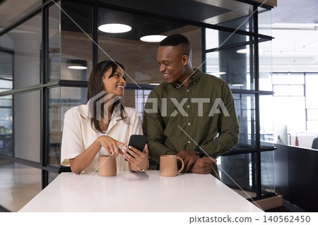 Diverse coworkers pointing at smartphone, leaning on white countertop in office break area Diverse coworkers pointing at smartphone, leaning on white countertop in office break area 140562450