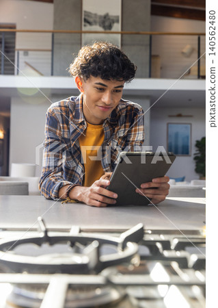 Man leaning on countertop holding tablet above gas stovetop with artwork, balcony, plant Man leaning on countertop holding tablet above gas stovetop with artwork, balcony, plant 140562480