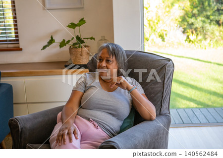Senior african american woman sitting in armchair with planter by sliding glass door Senior african american woman sitting in armchair with planter by sliding glass door 140562484