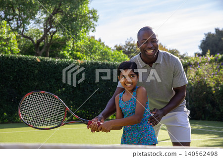 Diverse father and child guiding hand placement on tennis racket near net at backyard court Diverse father and child guiding hand placement on tennis racket near net at backyard court 140562498