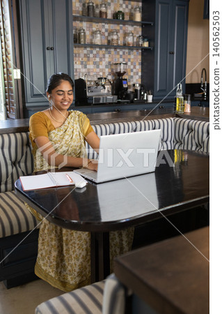 Indian woman in sari typing on laptop with wireless mouse, notebook at home kitchen nook 140562503