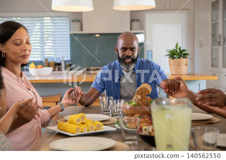Diverse family holding hands around dining table in modern kitchen with lemonade pitcher Diverse family holding hands around dining table in modern kitchen with lemonade pitcher 140562550