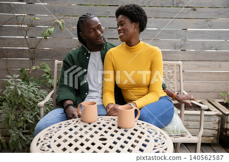 African american couple sitting and chatting on wooden deck around metal table holding coffee mugs African american couple sitting and chatting on wooden deck around metal table holding coffee mugs 140562587