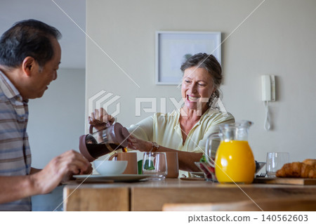 Diverse senior couple sitting at dining table in home area pouring coffee from french press Diverse senior couple sitting at dining table in home area pouring coffee from french press 140562603