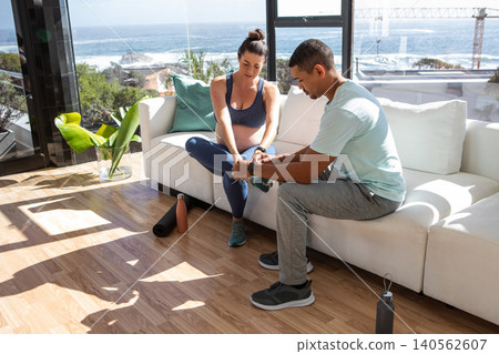 African American trainer and pregnant woman client tying running shoe on white sofa in living room African American trainer and pregnant woman client tying running shoe on white sofa in living room 140562607