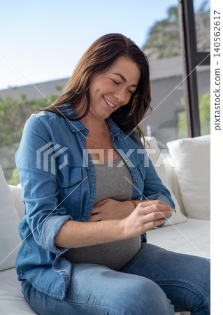 Mid adult expectant mother cradling belly while holding prenatal vitamin on sofa in living room Mid adult expectant mother cradling belly while holding prenatal vitamin on sofa in living room 140562617