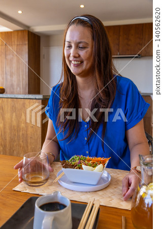 Asian woman sitting at dining table in kitchen with glass tumbler and smiling at colorful salad Asian woman sitting at dining table in kitchen with glass tumbler and smiling at colorful salad 140562620