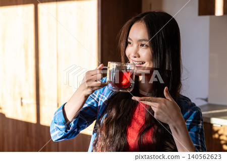 Asian woman pointing at double-walled glass mug filled with red tea in sunlit kitchen Asian woman pointing at double-walled glass mug filled with red tea in sunlit kitchen 140562623