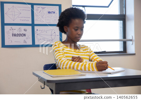 African american teenage girl writing in notebook at school desk with folder, math posters African american teenage girl writing in notebook at school desk with folder, math posters 140562655