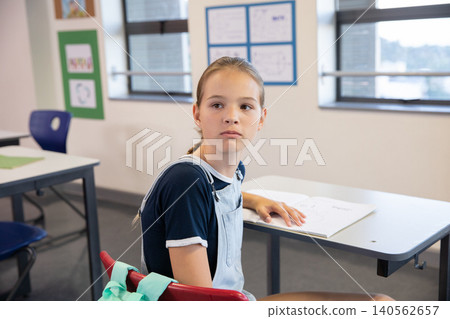 Adolescent student sitting at desk reviewing open workbook at school with red chair and jacket Adolescent student sitting at desk reviewing open workbook at school with red chair and jacket 140562657