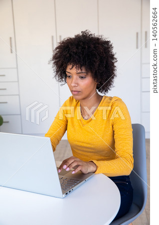 African american woman working on laptop at home nook by white cabinets in yellow top African american woman working on laptop at home nook by white cabinets in yellow top 140562664