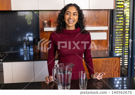Indian woman wearing burgundy sweater smiling behind kitchen counter with pitcher, empty glass Indian woman wearing burgundy sweater smiling behind kitchen counter with pitcher, empty glass 140562678