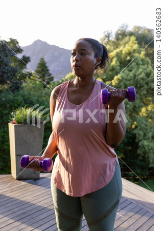 African american woman lifting purple dumbbells on backyard deck near planter under mountain sky African american woman lifting purple dumbbells on backyard deck near planter under mountain sky 140562683