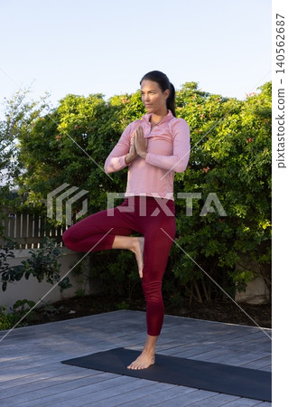 Woman practicing yoga in tree pose wearing pink top, red leggings on mat on deck Woman practicing yoga in tree pose wearing pink top, red leggings on mat on deck 140562687