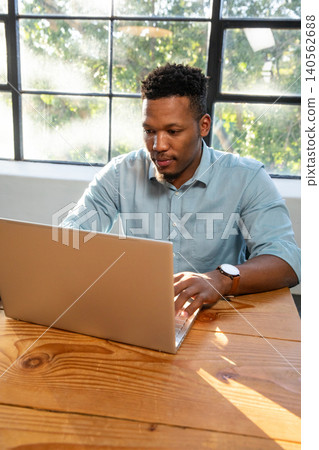 African american man typing on silver laptop at office table by grid window under sunlight African american man typing on silver laptop at office table by grid window under sunlight 140562688