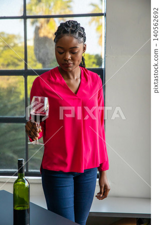 African american woman holding red wine glass near green bottle on dark tabletop by window African american woman holding red wine glass near green bottle on dark tabletop by window 140562692