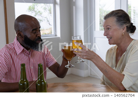 Senior couple clinking beer glasses while sitting at dining table with green bottles 140562704