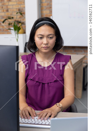 Asian woman typing on keyboard with desktop monitor and plant at office desk Asian woman typing on keyboard with desktop monitor and plant at office desk 140562711