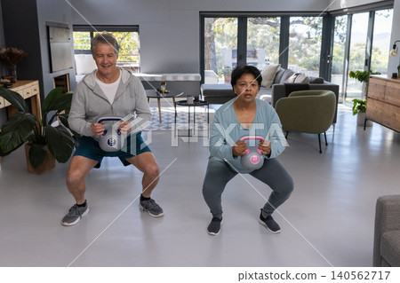 Diverse workout partners squatting with kettlebells in modern living room by glass doors 140562717