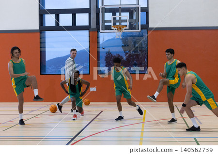 Basketball coach along with five players in green jerseys performing lunges under hoop Basketball coach along with five players in green jerseys performing lunges under hoop 140562739