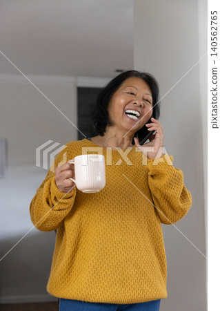 Senior asian woman talking on smartphone with ceramic mug, smiling inside living room Senior asian woman talking on smartphone with ceramic mug, smiling inside living room 140562765
