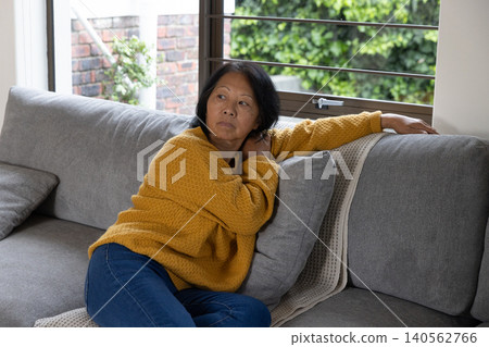 Senior asian woman in mustard sweater, jeans reclining on sofa cushion gazing through blinds Senior asian woman in mustard sweater, jeans reclining on sofa cushion gazing through blinds 140562766