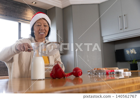 Senior Asian woman wearing Santa hat sifting flour through metal sieve into bowl at kitchen counter Senior Asian woman wearing Santa hat sifting flour through metal sieve into bowl at kitchen counter 140562767