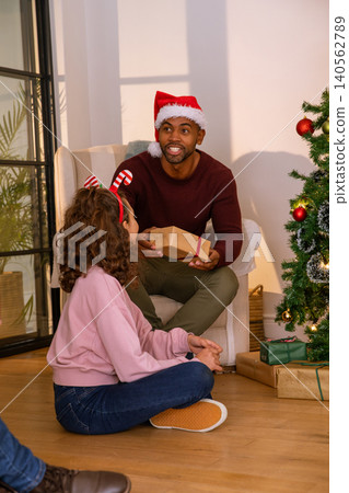 Diverse father wearing santa hat and daughter exchanging gift beside christmas tree in living room Diverse father wearing santa hat and daughter exchanging gift beside christmas tree in living room 140562789