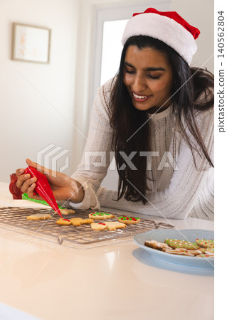 Asian woman wearing Santa hat piping red icing onto sugar cookies on cooling rack in kitchen Asian woman wearing Santa hat piping red icing onto sugar cookies on cooling rack in kitchen 140562804
