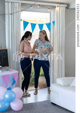 Diverse female friends standing by sliding door at home with swaddled baby under pennant bunting Diverse female friends standing by sliding door at home with swaddled baby under pennant bunting 140562813