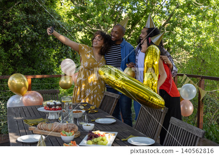 Diverse friends wearing party hats taking selfie on deck with smartphone by table near balloons 140562816