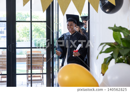 Classmates entering living room wearing black gowns and holding diploma with ribbon Classmates entering living room wearing black gowns and holding diploma with ribbon 140562825