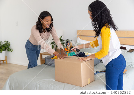 Diverse mom and teenage daughter sealing and packing cardboard box in bedroom beside suitcase Diverse mom and teenage daughter sealing and packing cardboard box in bedroom beside suitcase 140562852