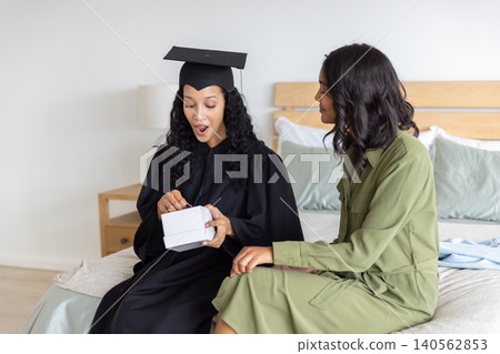 Diverse mom and teenage daughter opening white gift box in bedroom wearing graduation gown Diverse mom and teenage daughter opening white gift box in bedroom wearing graduation gown 140562853