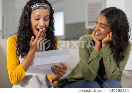 Diverse mom and teenage daughter sitting on couch in living room reading letter smiling Diverse mom and teenage daughter sitting on couch in living room reading letter smiling 140562854