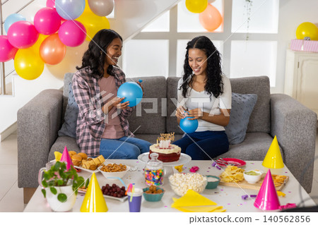 Diverse mom and teenage daughter inflating bright blue balloons in living room with birthday cake Diverse mom and teenage daughter inflating bright blue balloons in living room with birthday cake 140562856